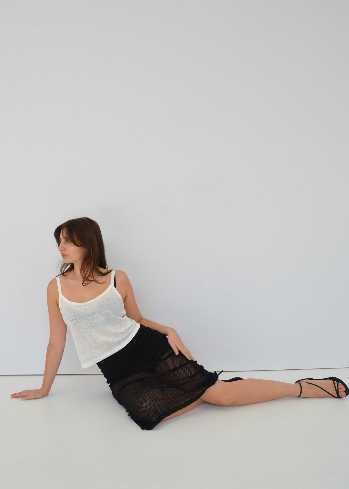 Woman sitting on a white floor wearing a white japanese linen cami top and black italian lace skirt against a white wall.