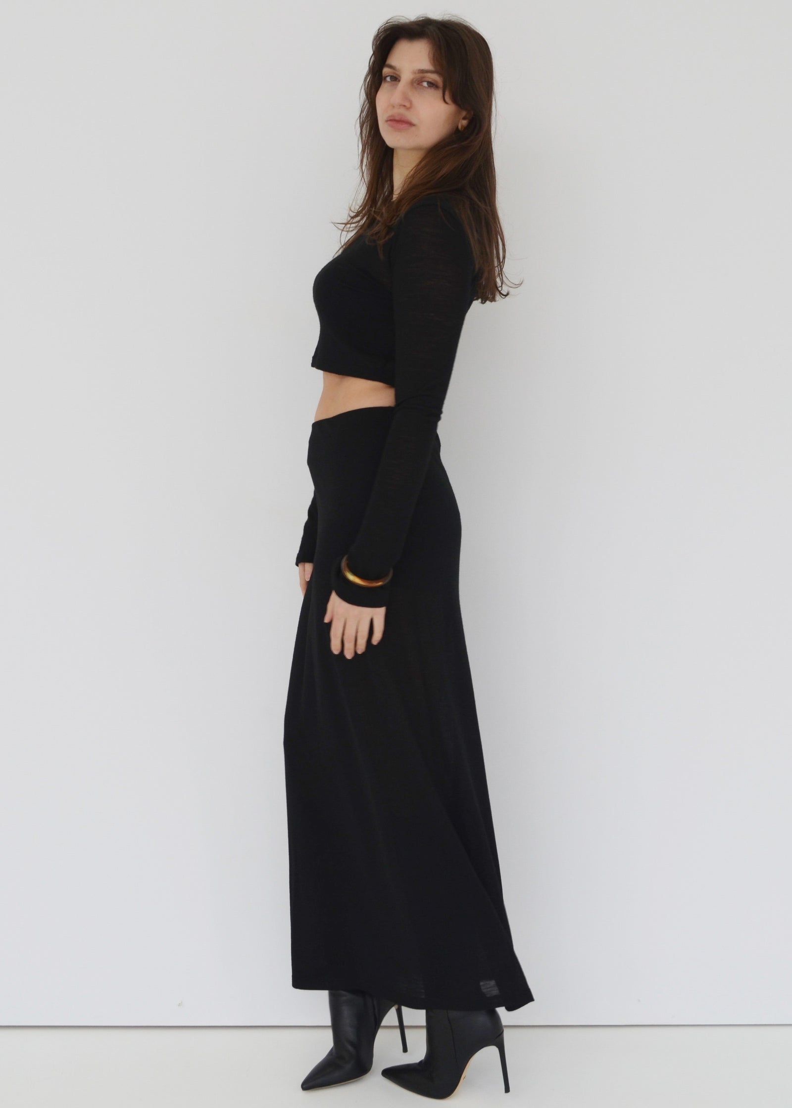 Woman facing the side wearing the maxi Isabel skirt and the matching Tati flared long sleeve crop top in sheer black Japanese Tencel and merino knit against a white wall. Australian made. 