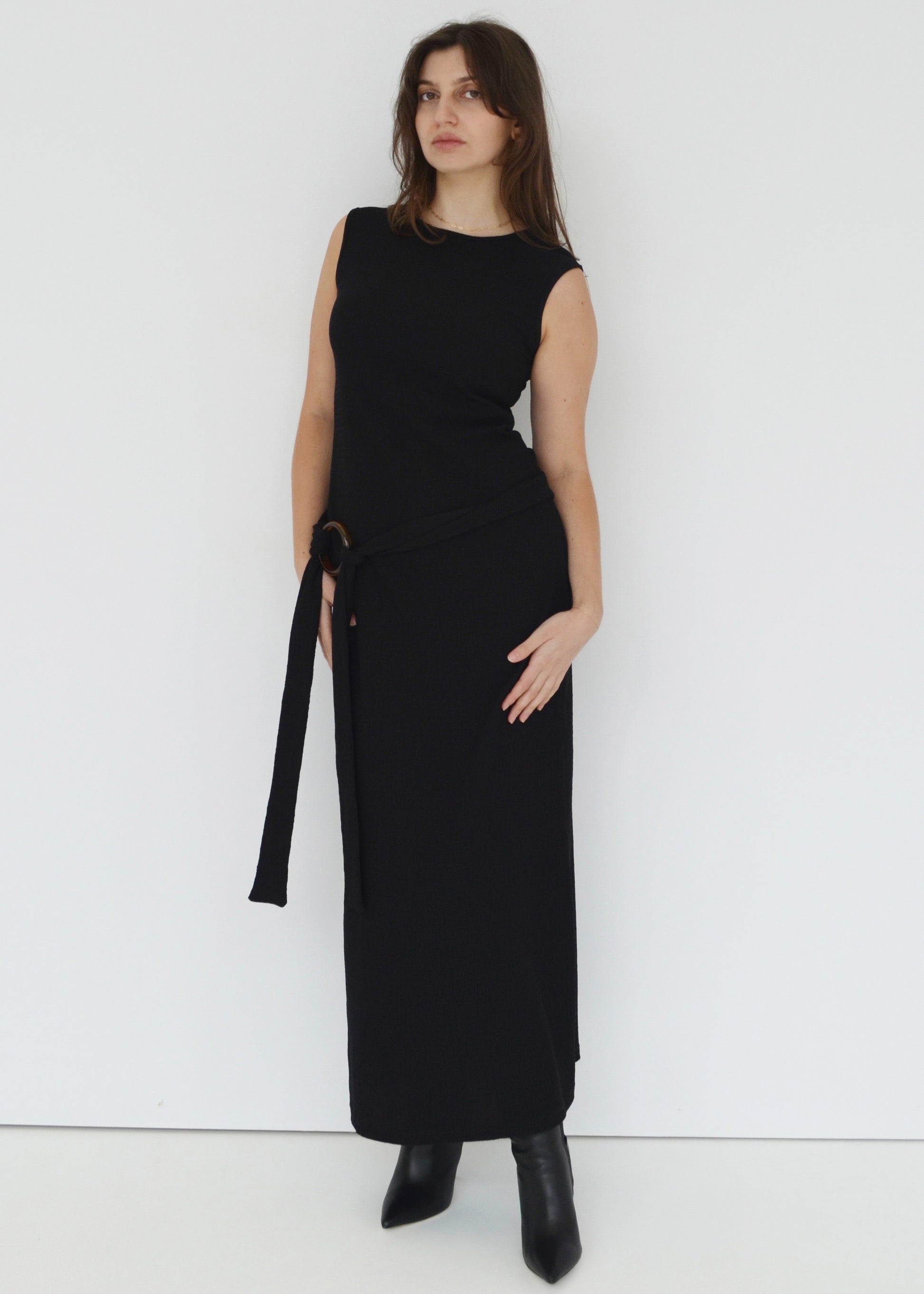 Woman wearing the sleeveless Domenica maxi dress in textured black shirred crinkle fabric with tortoise shell hoop and tie belt around waist against white wall. Australian made. 