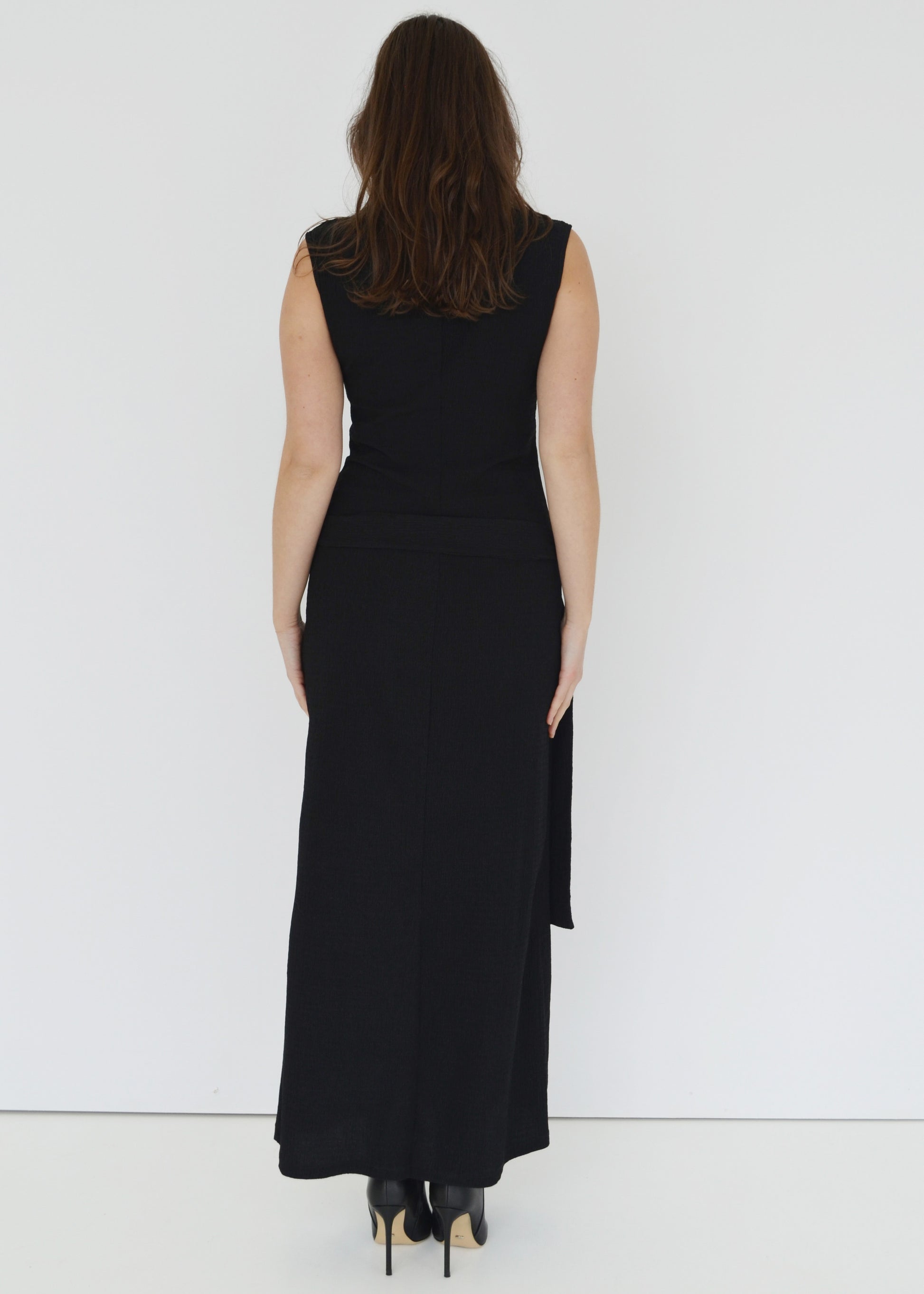 Woman facing the back side wearing the sleeveless Domenica maxi dress in textured black shirred crinkle fabric with tie belt and tortoise shell hoop around waist against white wall. Australian made. 