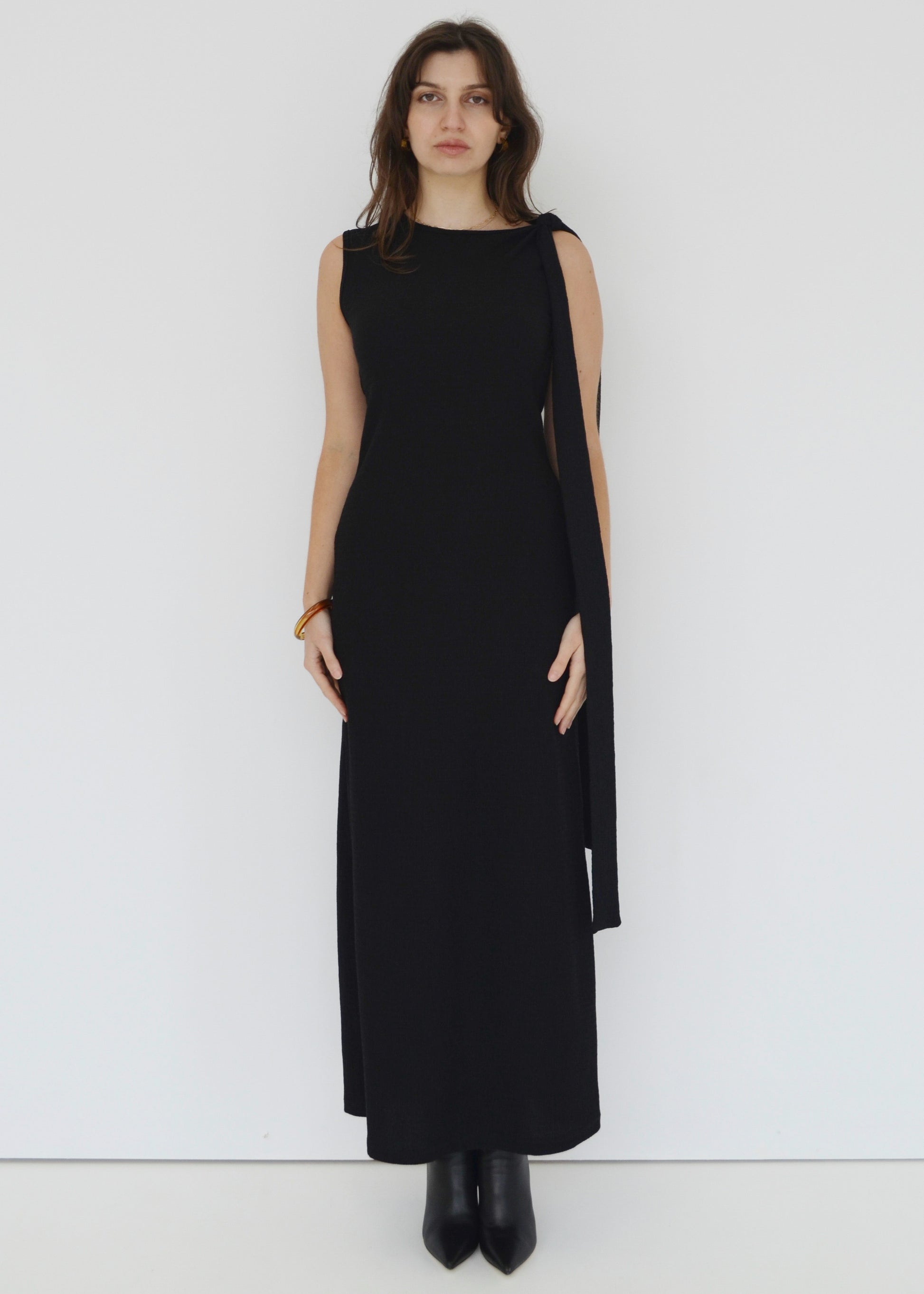 Woman facing front wearing the sleeveless Domenica maxi dress in textured black shirred crinkle fabric with tie belt tied around shoulder against white wall. Australian made. 