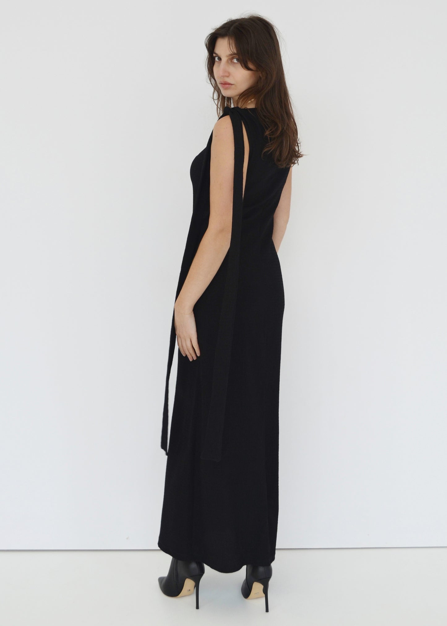 Woman facing the back side wearing the sleeveless Domenica dress in textured black shirred crinkle fabric with tie belt tied around shoulder against white wall. Australian made. 