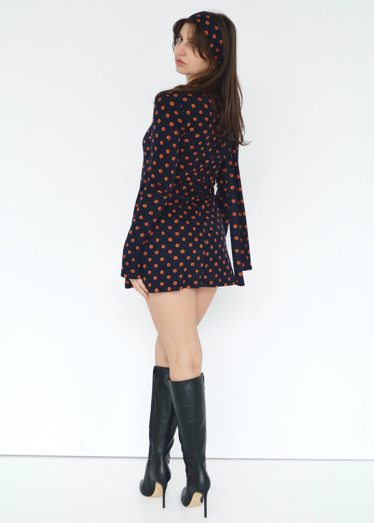 Woman wearing a black dress with red & yellow polka dots and black knee-high boots on a white background back side