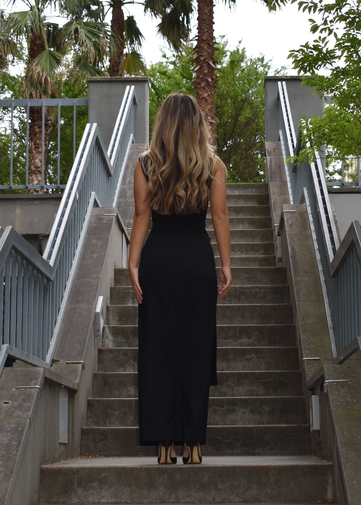 Woman facing the back side wearing the sleeveless Domenica maxi dress in textured black shirred crinkle fabric with tie belt around waist on staircase. Australian made. 