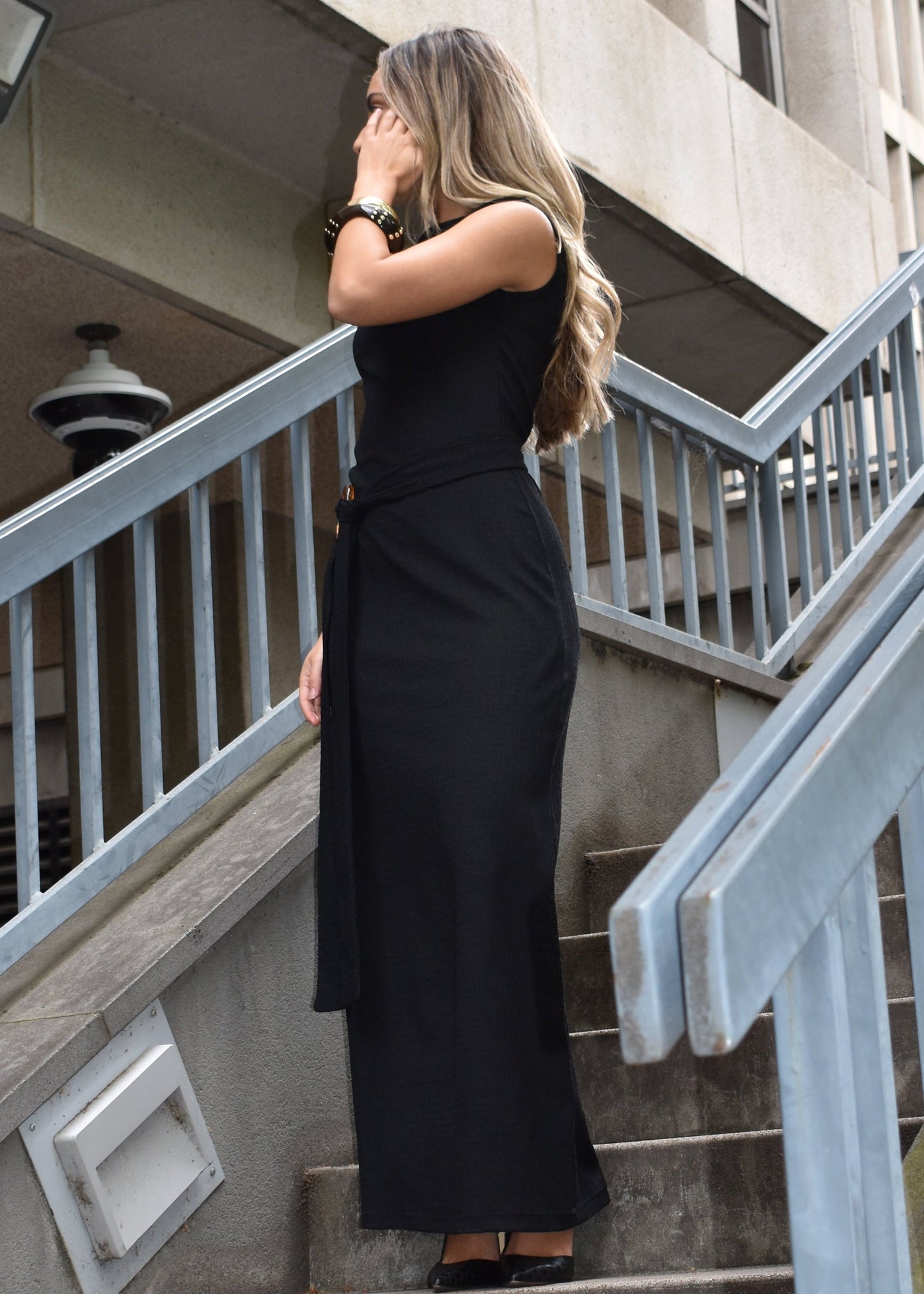 Woman wearing the Domenica maxi dress in textured black shirred crinkle fabric with tortoise shell hoop and tie belt around waist on staircase. Australian made. 