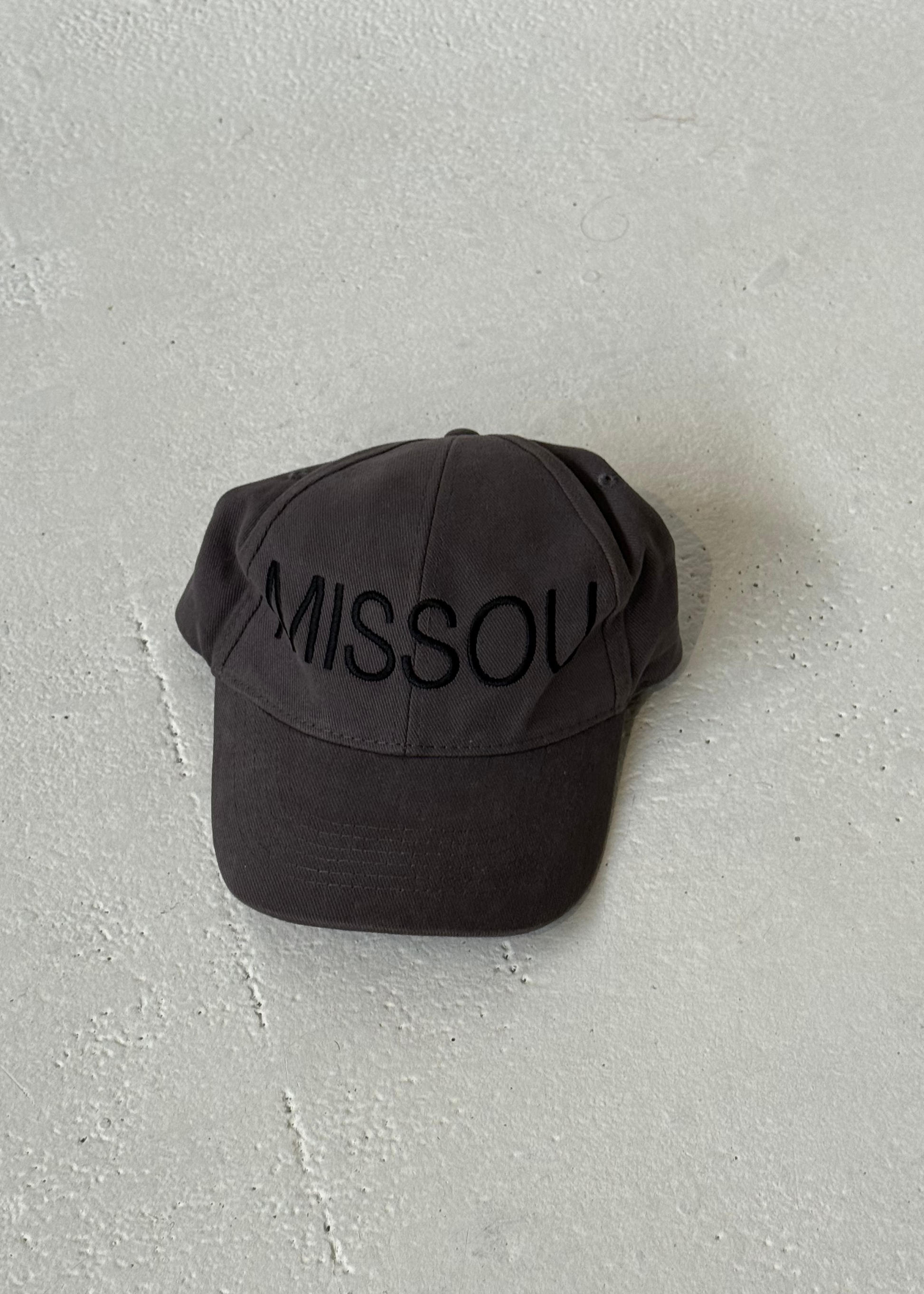 Front of the Missou cap in charcoal grey cotton with embroidered block logo on a stone floor. 
