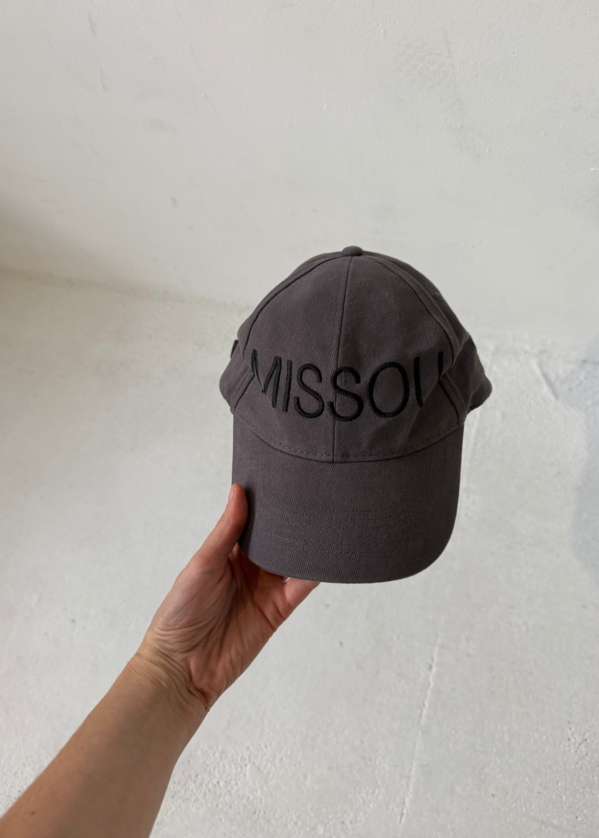 Woman holding the Missou cap in charcoal grey cotton with embroidered block logo against a stone wall. 