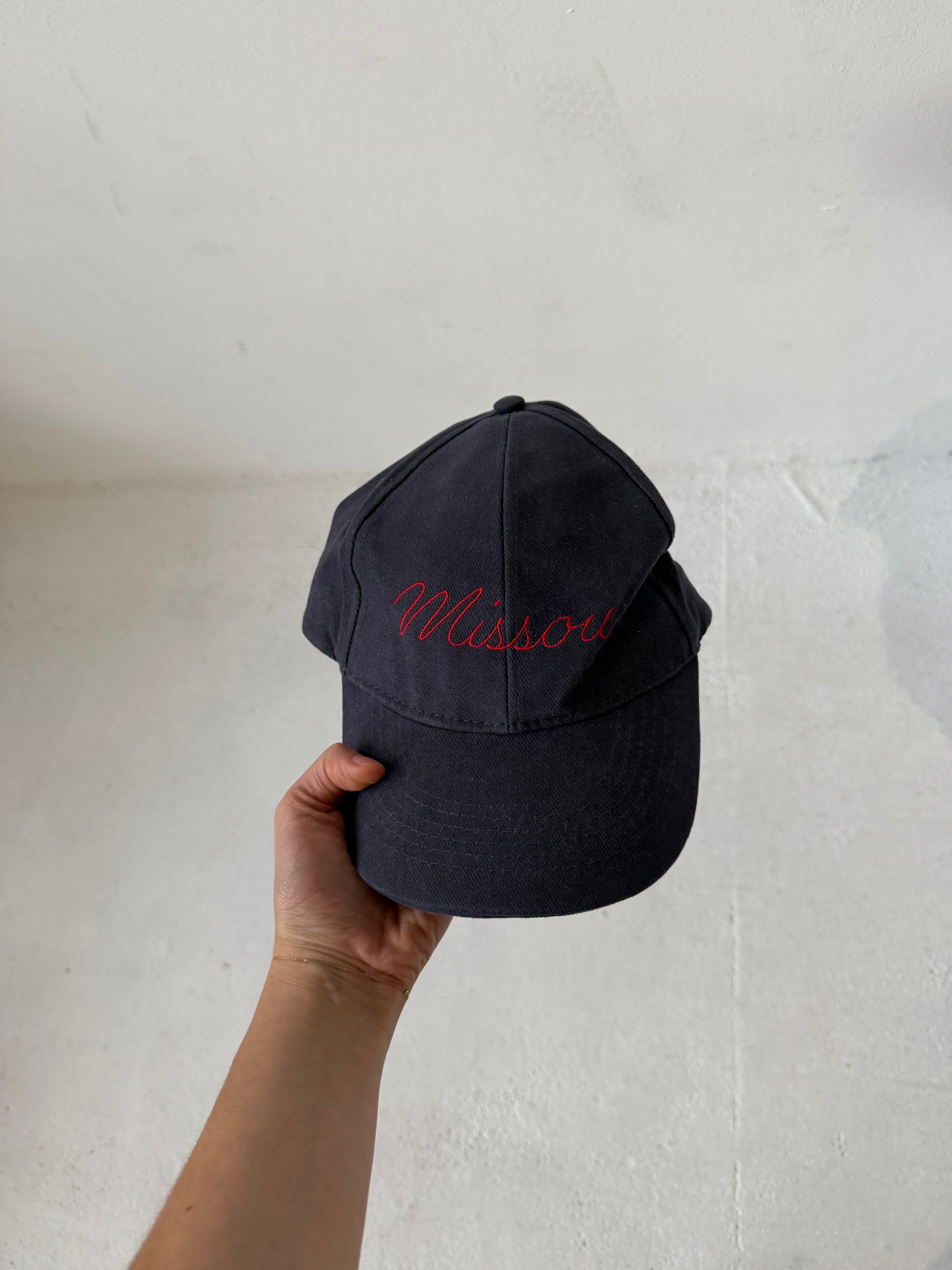 Woman holding the Missou cap in navy cotton with red embroidered cursive logo in front of a stone wall. 