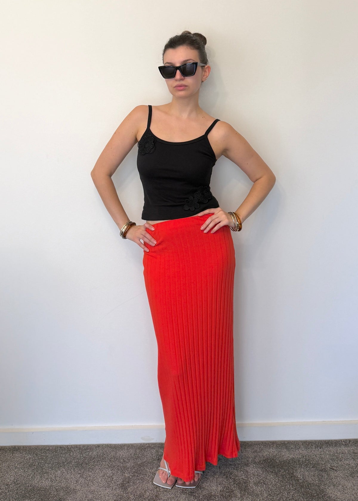 A woman stands against an off-white wall, wearing a Missou black flower applique Toto tank top, Missou Alba ribbed maxi skirt, and bracelets. 