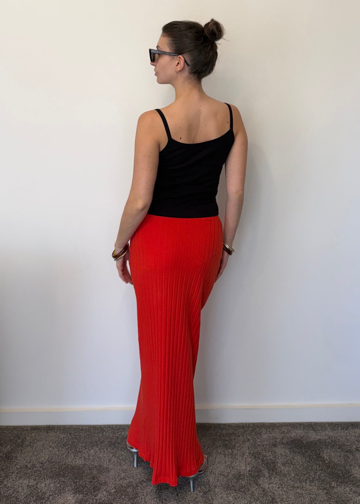 A woman stands against an off-white wall, wearing a Missou black flower applique Toto tank top, Missou Alba ribbed maxi skirt, and bracelets. 
