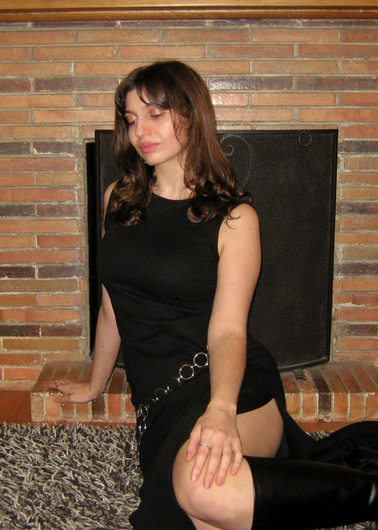 A woman wearing the Missou Celia black sleeveless maxi dress and black boots with the Missou M silver chain belt sits in front of a brick fireplace.