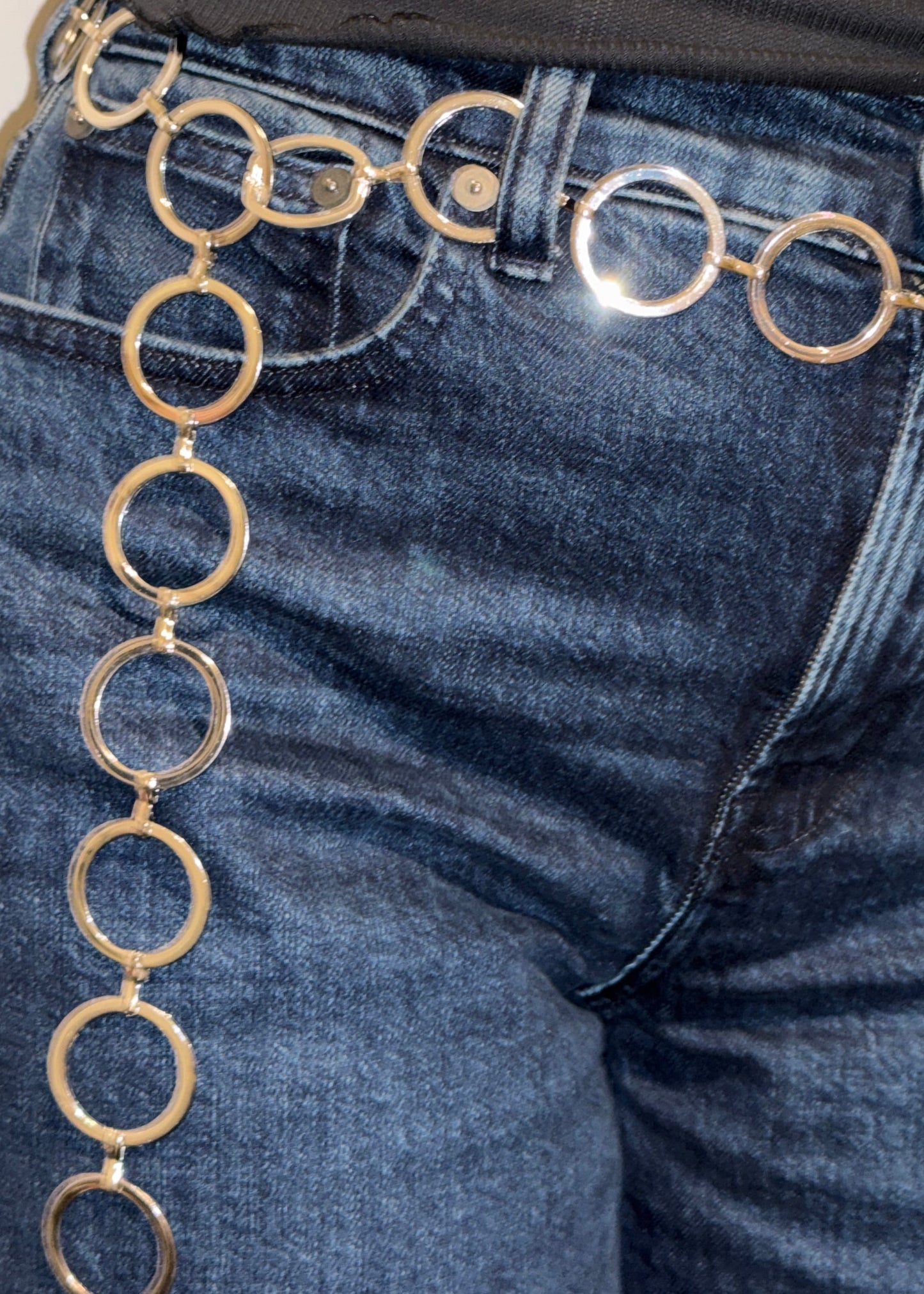 A close up of a woman wearing dark blue denim jeans and a black ribbed top, with a metallic silver chain belt featuring large circular links draped across the waistband and down the side.