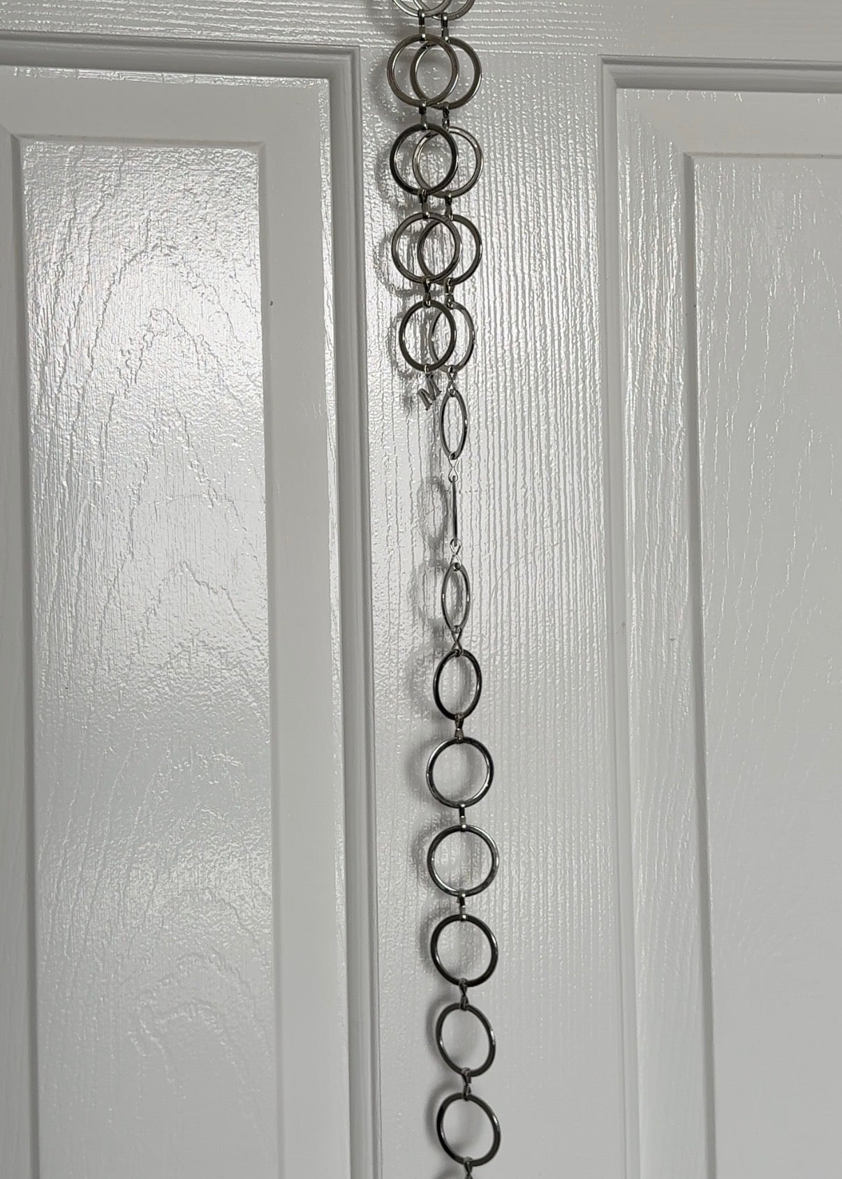 A vertical silver metal chain belt made of interlocking circular links hanging down the center of a white paneled door.