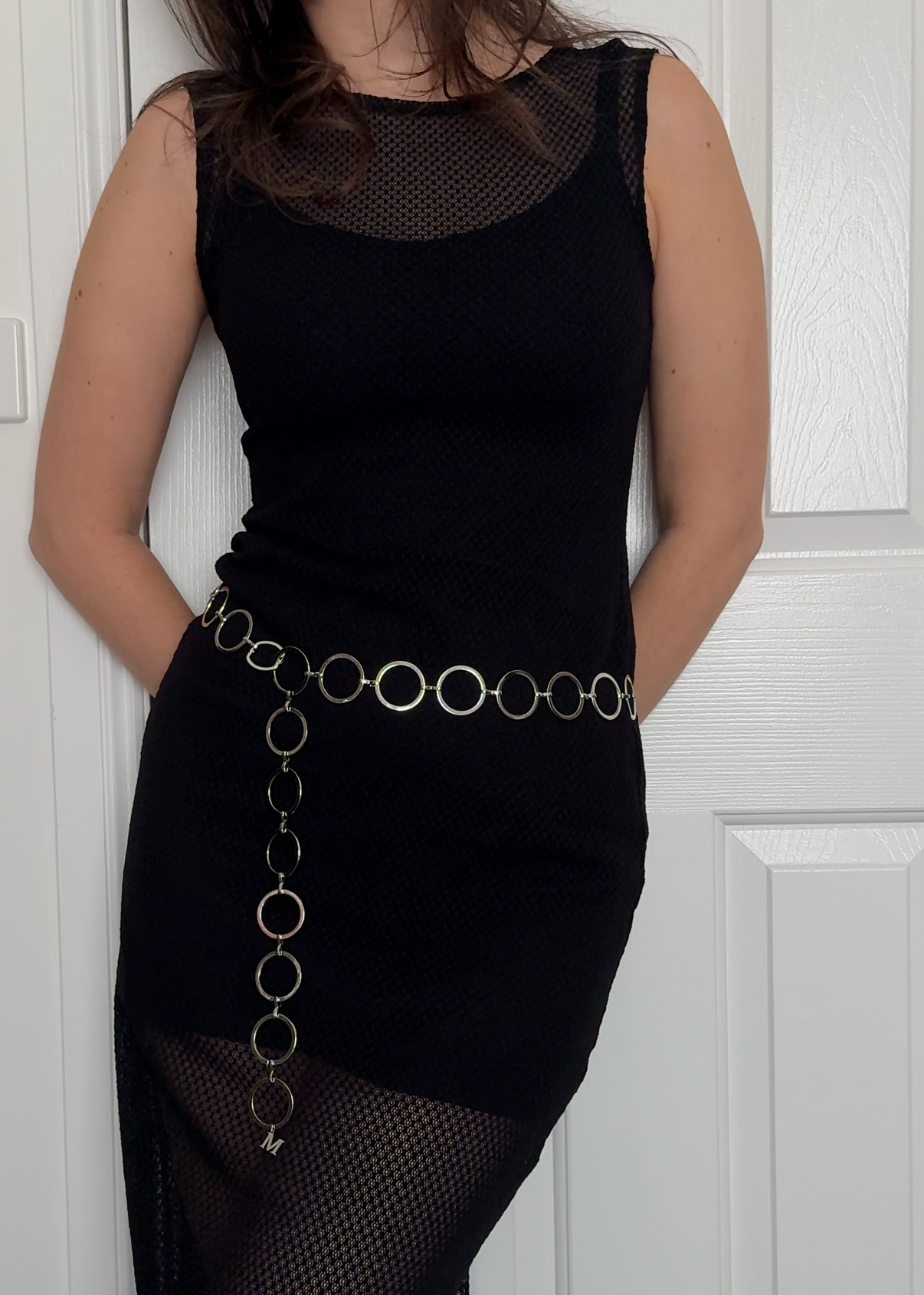 A woman wearing a sleeveless black Italian lace Missou dress and a belt made of large interlocking metal rings and an M pendent, standing against a white door.
