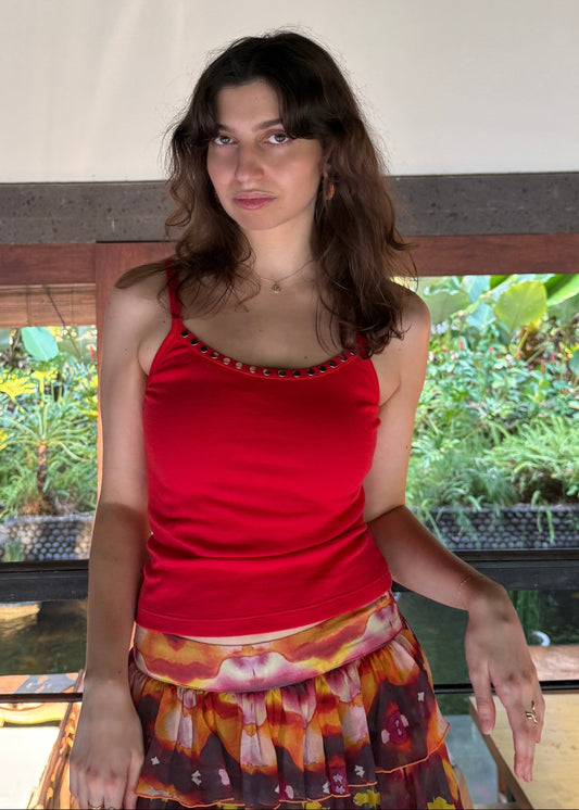 A woman wearing a Missou red Toto tank top and a colorful patterned skirt stands on an outdoor balcony with a view of green plants below.
