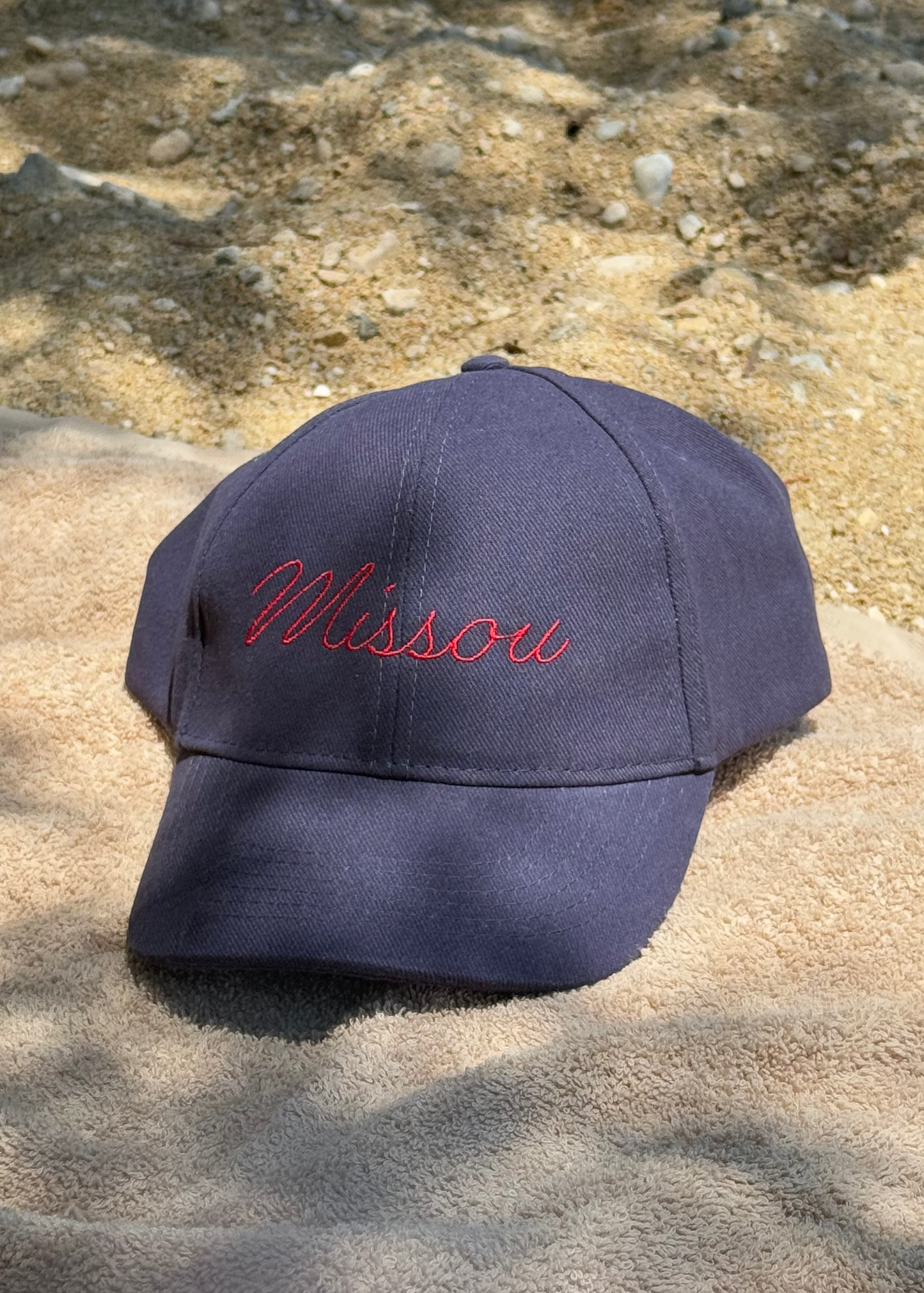 The Missou cap in navy cotton with red embroidered cursive logo towel at a beach in Greece with the sun shining through the trees and sand and shells in the background. 