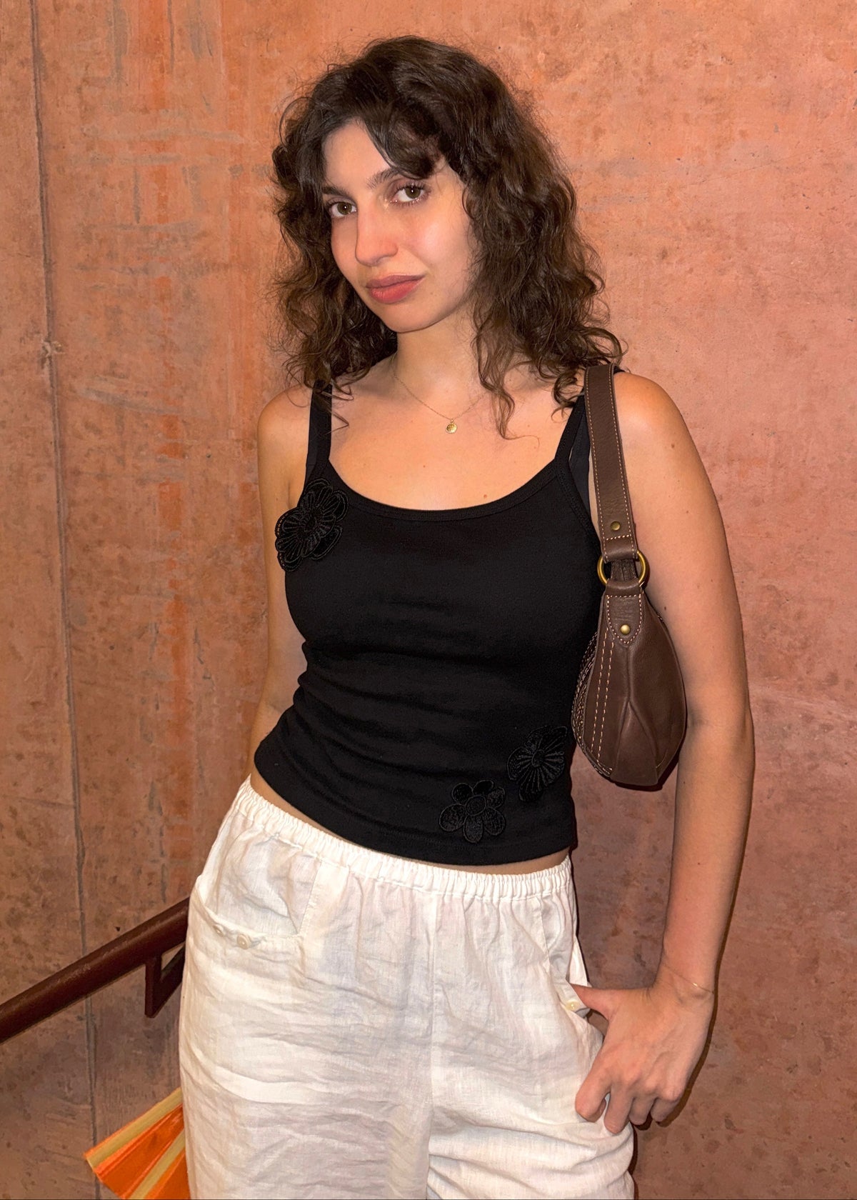 A woman stands against textured terracotta wall, wearing a Missou black flower applique Toto tank top, white pants, and carrying a brown shoulder bag.
