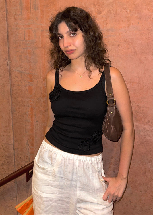 A woman stands against textured terracotta wall, wearing a Missou black flower applique Toto tank top, white pants, and carrying a brown shoulder bag.